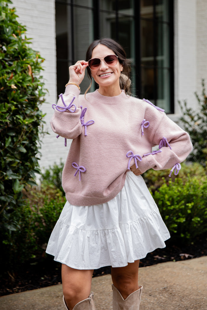 Woman wearing a pink sweater with purple bows and a white skirt outdoors.