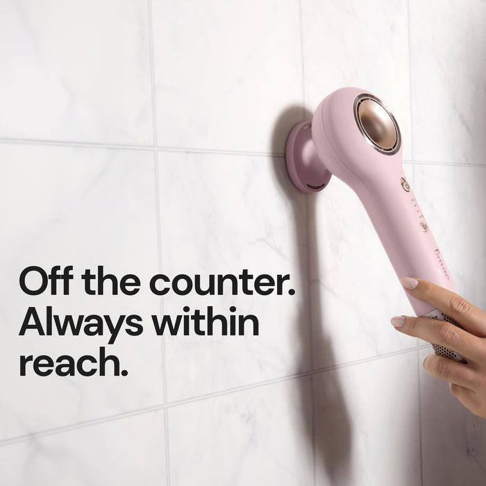 Pink L'ange hair dryer attached to a magnetic wall mount on marble tile, held by a hand, with text "Off the counter. Always within reach."