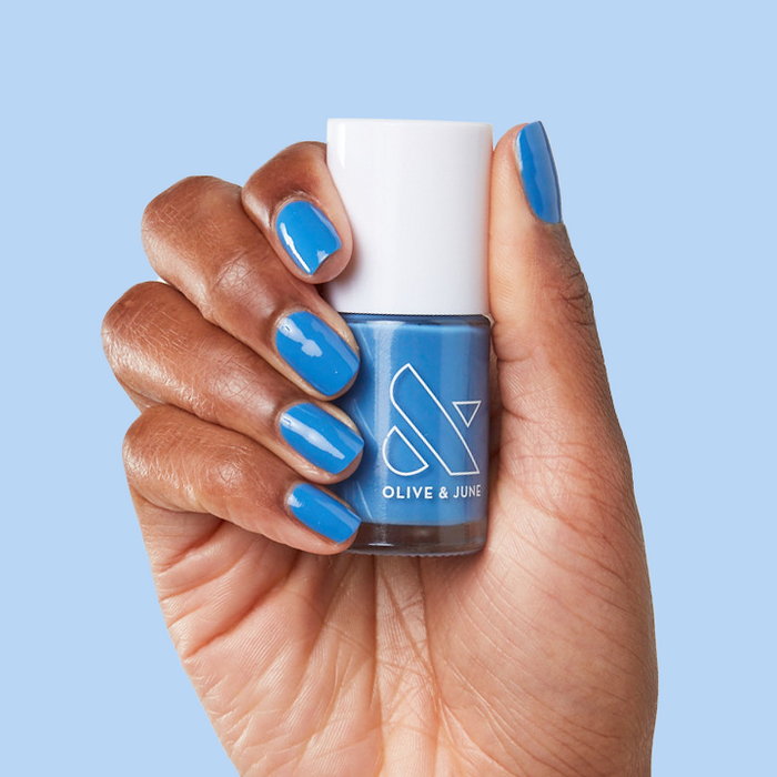 Hand with nails painted in Blue Canoe long-lasting polish holding Olive and June polish bottle against blue background