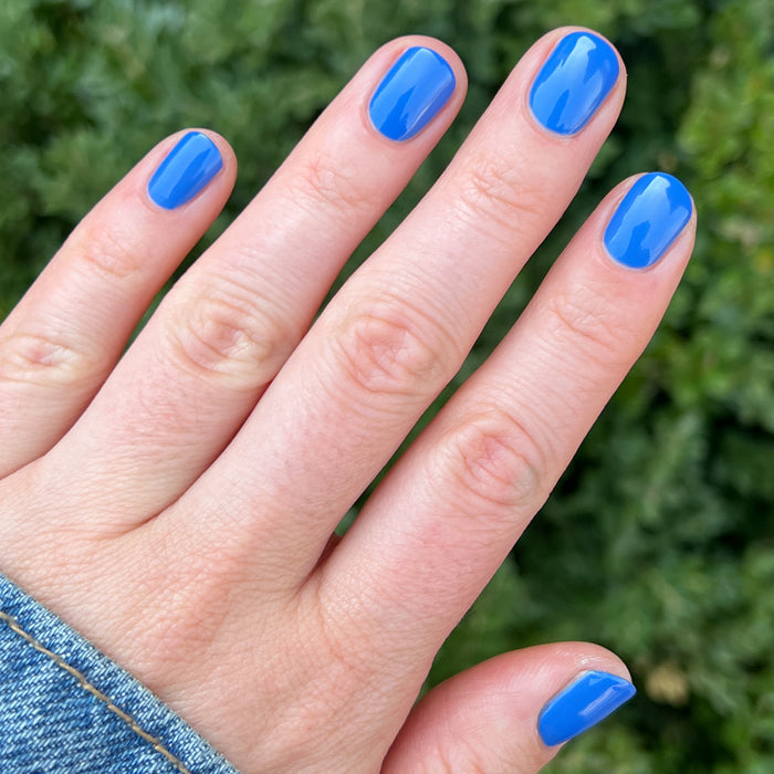Hand with short nails painted in Blue Canoe gel polish by Olive and June against outdoor greenery background