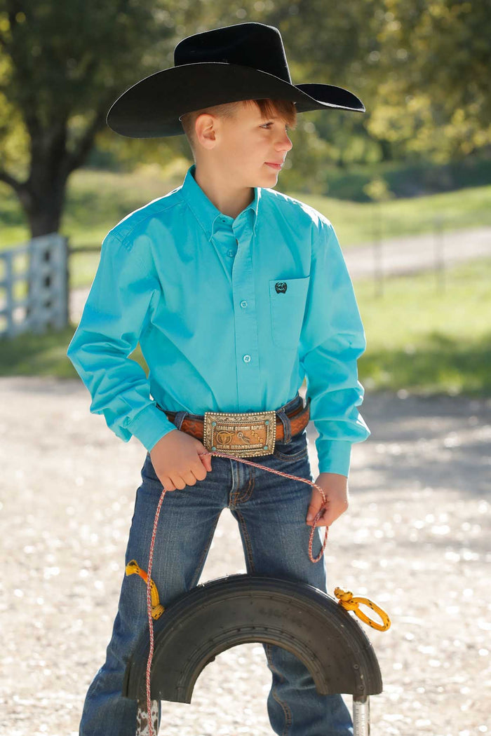 A boy wearing a turquoise long sleeve button down shirt with an embroidered logo on the pocket.