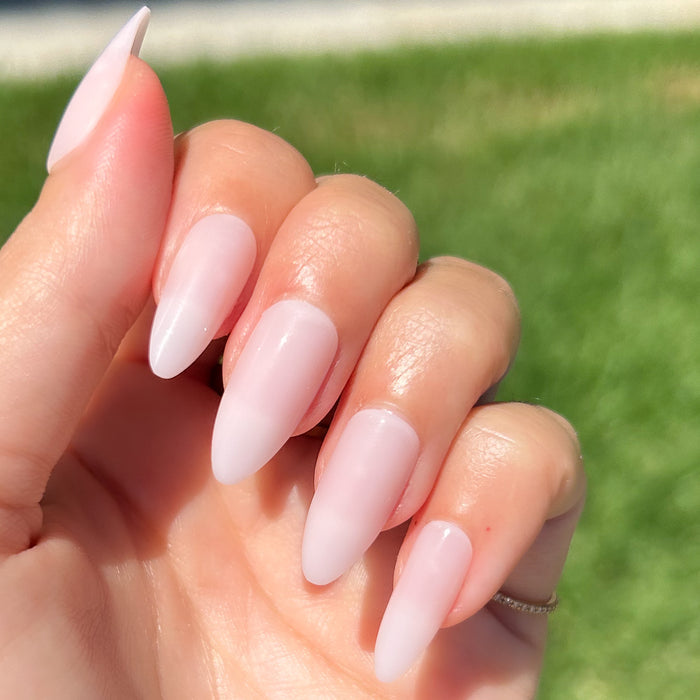CCT - Press-On | Long | Almond showing hand with sheer soft pink long almond press-on manicure outdoors on grass background