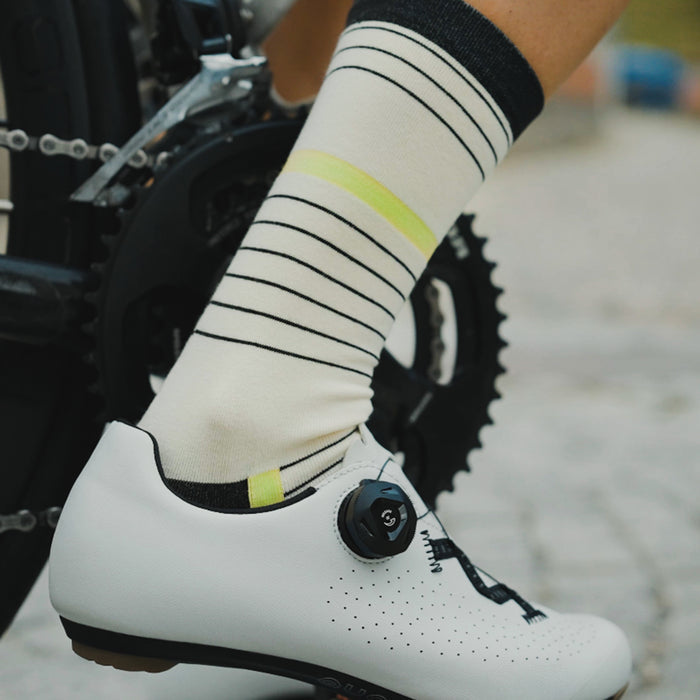 A close-up of a person’s foot wearing a white cycling shoe with a dial closure and cream-colored socks with black and yellow stripes, positioned next to a bicycle crank and chainring.