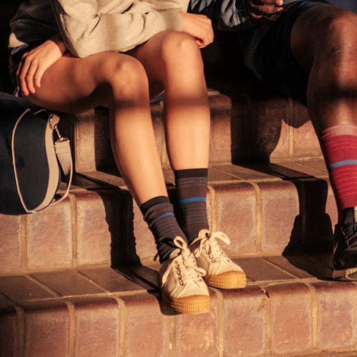 Two people sitting side by side on brick steps, with only their legs and part of their torsos visible. One person wears a gray sweatshirt, shorts, and striped socks with sneakers; the other wears darker clothing and striped socks.