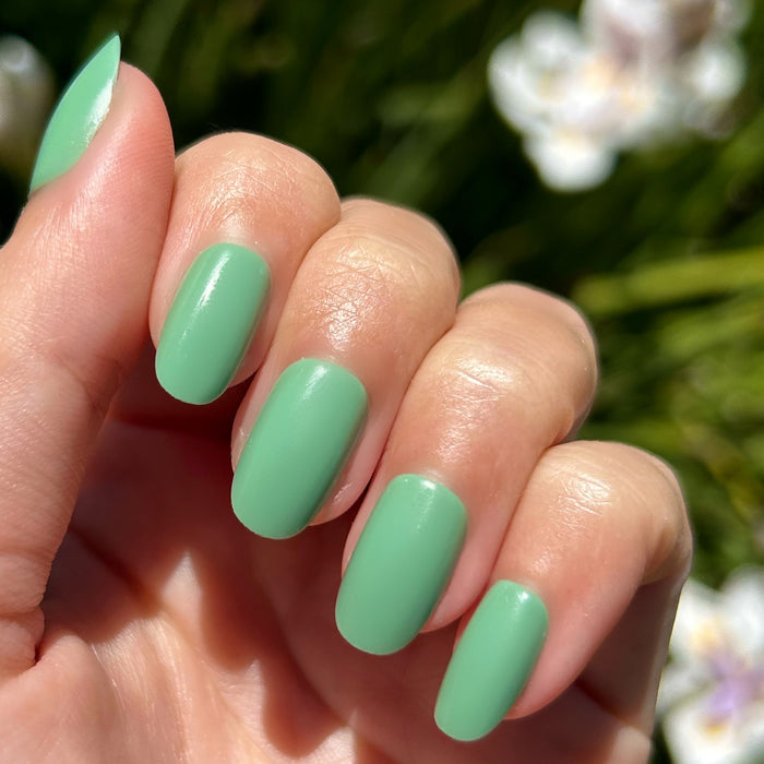 Cucumber Sandwich - Press-On | Short | Round showing hand with smooth short round green false nails outdoors with blurred flowers background