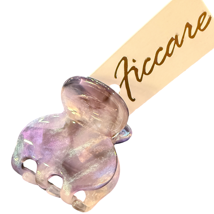 A close-up of a decorative hair claw clip with an iridescent lavender and pink elephant head charm, attached to a beige paper tag printed with the brand name 'Ficcare' in gold script, set against a plain white background.