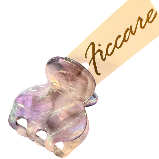A close-up of a decorative hair claw clip with an iridescent lavender and pink elephant head charm, attached to a beige paper tag printed with the brand name 'Ficcare' in gold script, set against a plain white background.