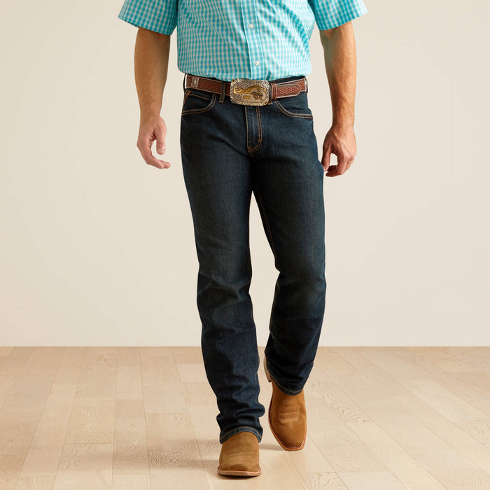 Men's Ariat M4 dark wash Pro series relaxed fit boot cut jean. Front view showing bottom of jeans stacked on boots.