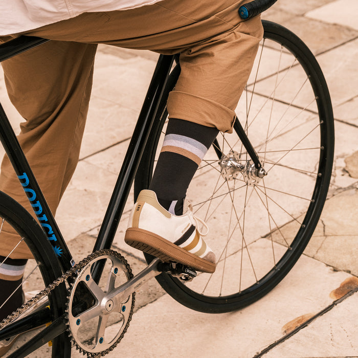 A person wearing tan pants, striped socks, and white sneakers rides a black bicycle on a light-colored stone pavement. Only the lower half of the body and part of the bike are visible.