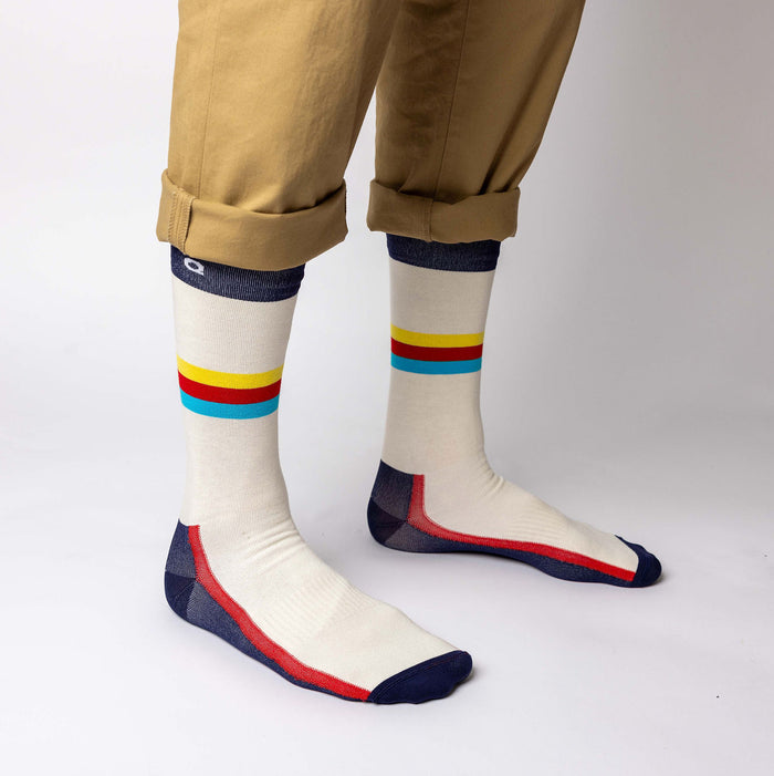 A person wearing tan pants with cuffs rolled up, showing off cream-colored socks with yellow, red, and blue stripes, and navy blue accents on the toes and heels, standing against a plain white background.
