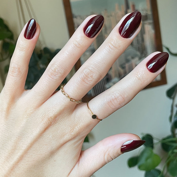 Obsessed - Press-On | Medium | Almond showing hand with deep dark ruby almond press-on manicure indoors with gold rings