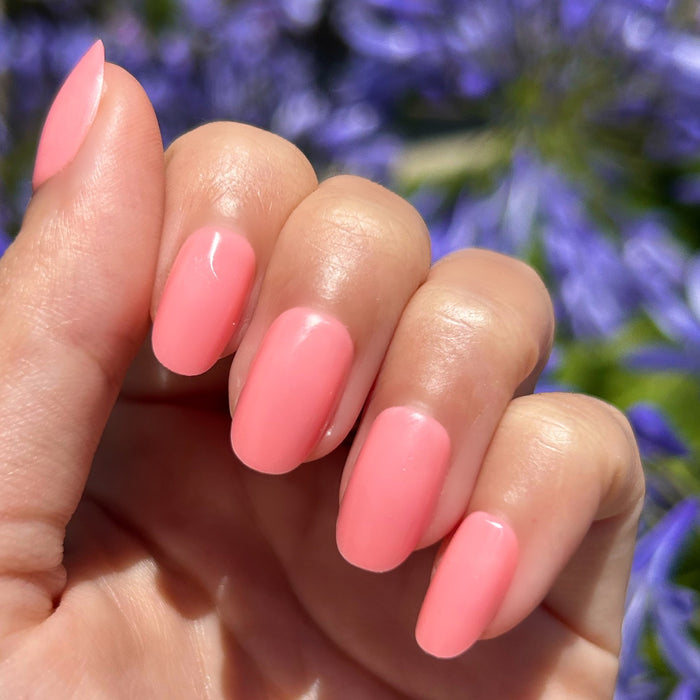 Pen Pal - Press-On | Short | Round showing hand with short round bright bubblegum pink false nails outdoors with purple flowers background
