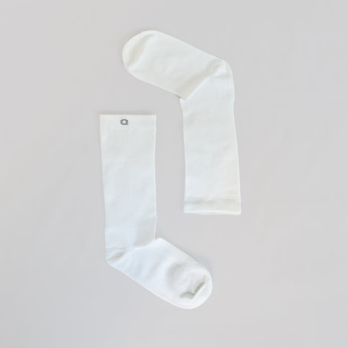 Two plain white socks laid flat on a light surface, positioned to form an angled shape resembling an upside-down letter L. One sock has a small gray circular emblem near the top.