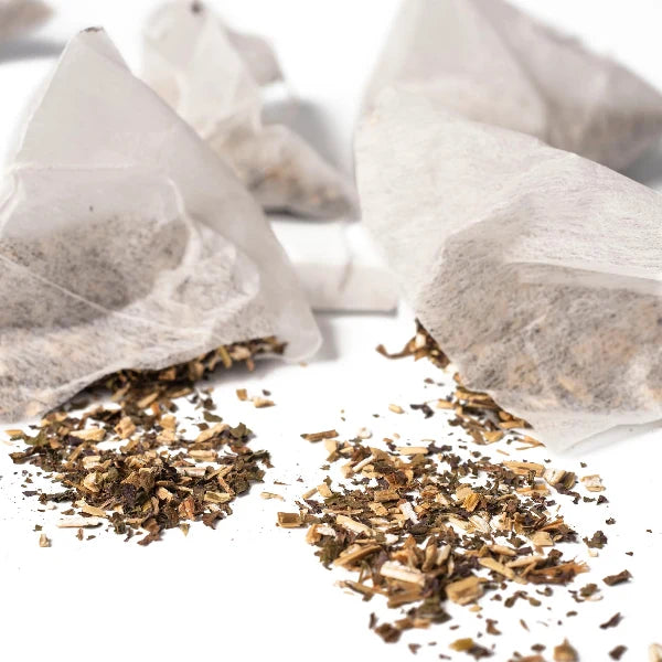 Several white paper teabags with loose-leaf tea spilled onto a white surface, showing the brown and tan tea leaves inside and scattered around.