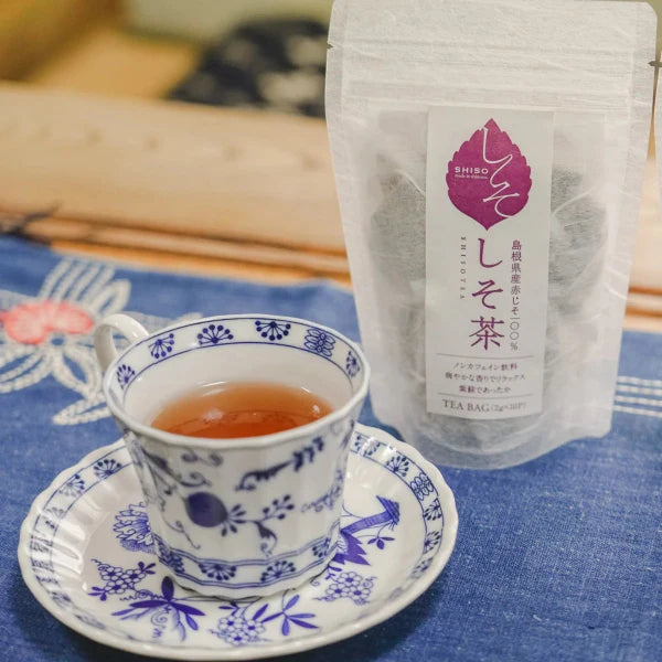 A white and blue floral ceramic teacup with a matching saucer filled with amber tea, standing beside a clear stand-up pouch of Japanese tea on a blue fabric surface.