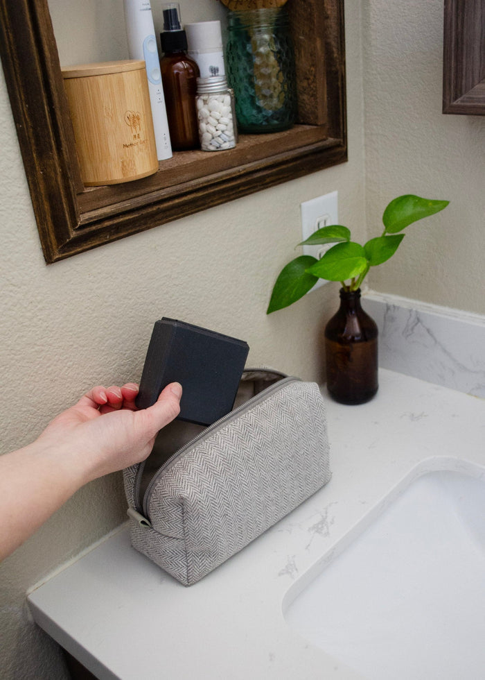 Person reaching for a toiletry bag in a gray pouch on a bathroom counter with a mirror and plants in the background.