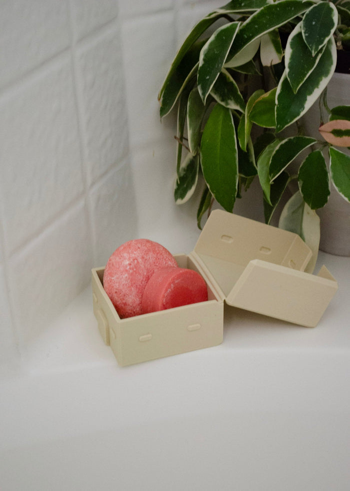 Pink shampoo and conditioner bar in a sand color silicone travel case on a white surface with a plant in the background