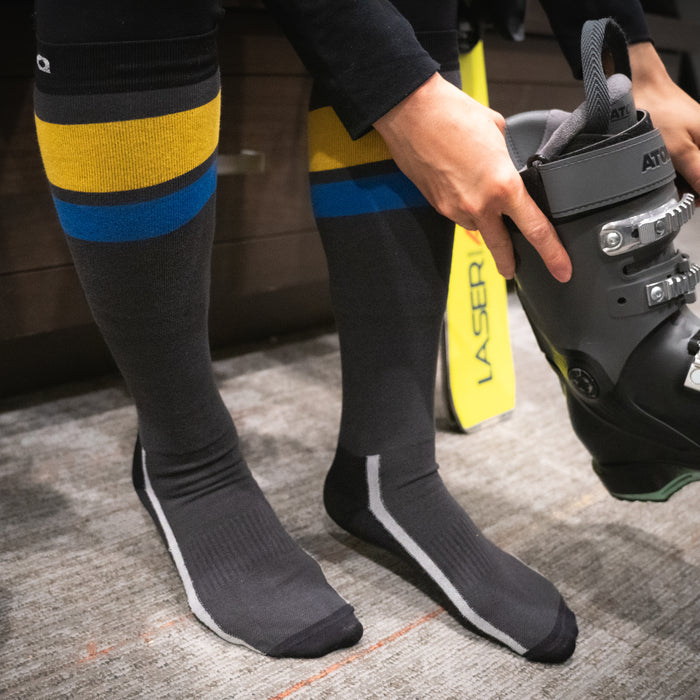 A person wearing black knee-high socks with blue and yellow stripes is putting on a black ski boot while sitting indoors on a carpeted floor.