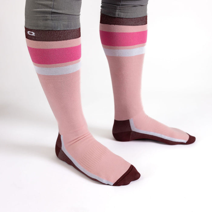 A person wearing knee-high socks with pink, maroon, and white stripes, standing on a white surface. Only the legs and feet are visible.