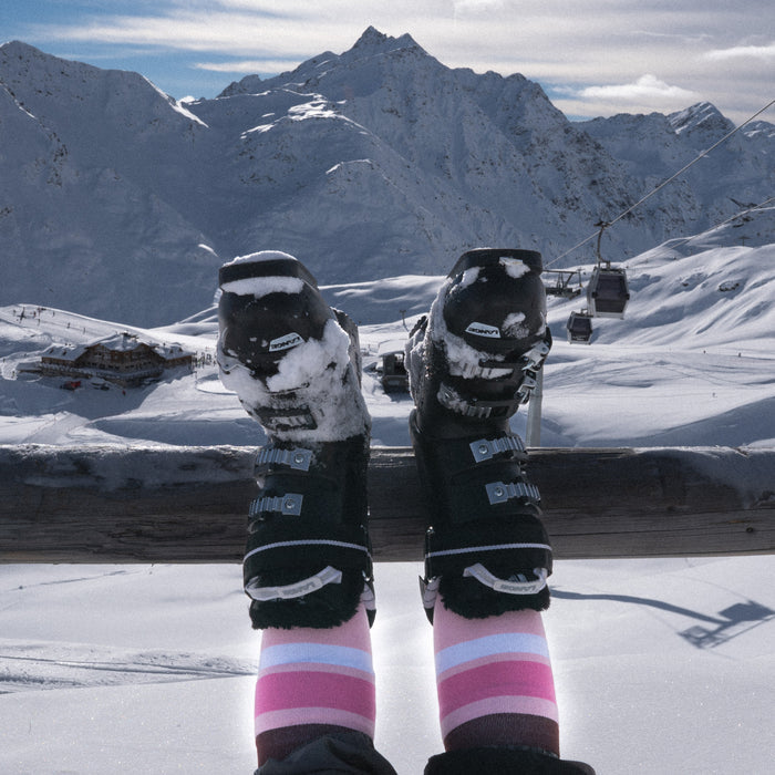 Person’s legs in striped socks and ski boots rest on a wooden railing with a snowy mountain landscape, ski lifts, and chalets in the background under a partly cloudy sky.
