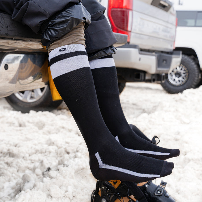 Two people wearing black knee-high socks with white stripes and crampons sit on a tailgate with their feet extended over snowy ground. A truck and another vehicle are visible in the background.
