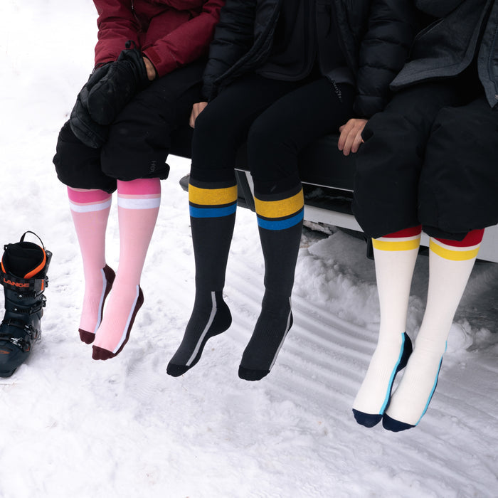 Three people sit side by side in the snow, wearing colorful knee-high socks with stripes. Their feet dangle above the ground, and a single black winter boot is visible next to them.