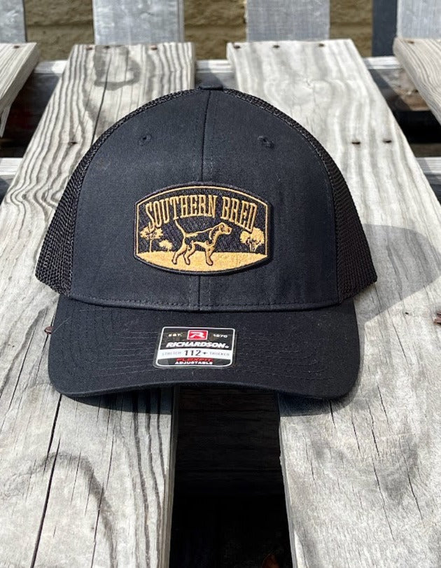 A black Southern Bred baseball cap with a logo on the front and an adjustable snapback closure, placed on a wooden surface.
