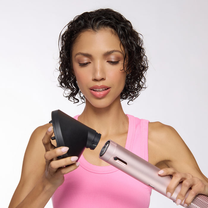 A woman in a pink tank top attaching a black angled diffuser to a rose-gold hair dryer (TurboStyle)